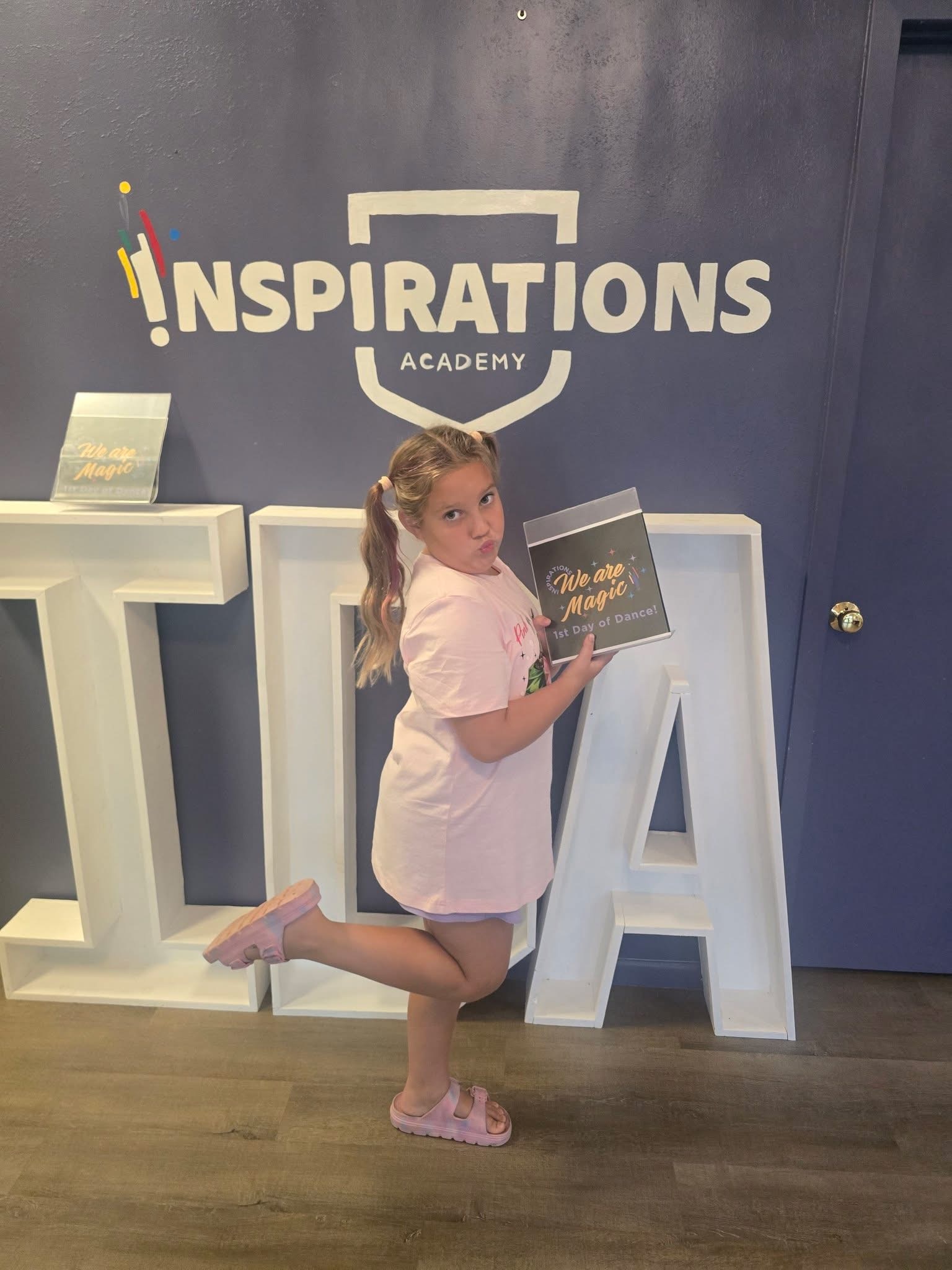 A young Inspirations Academy student holding a We Are Magic first day of dance sign in front of the studio wall logo