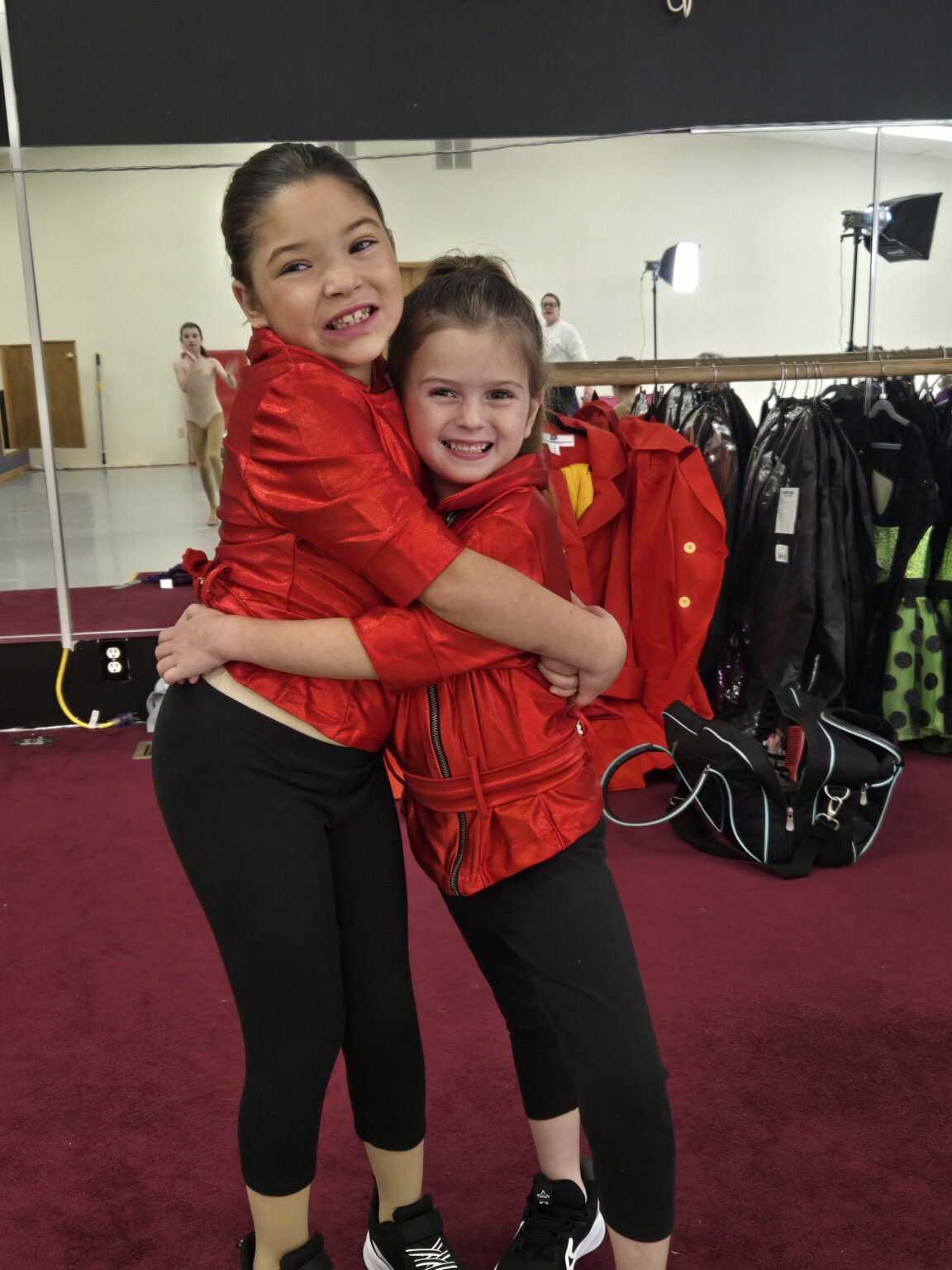 Two young dancers in red costumes hugging in the studio before a performance