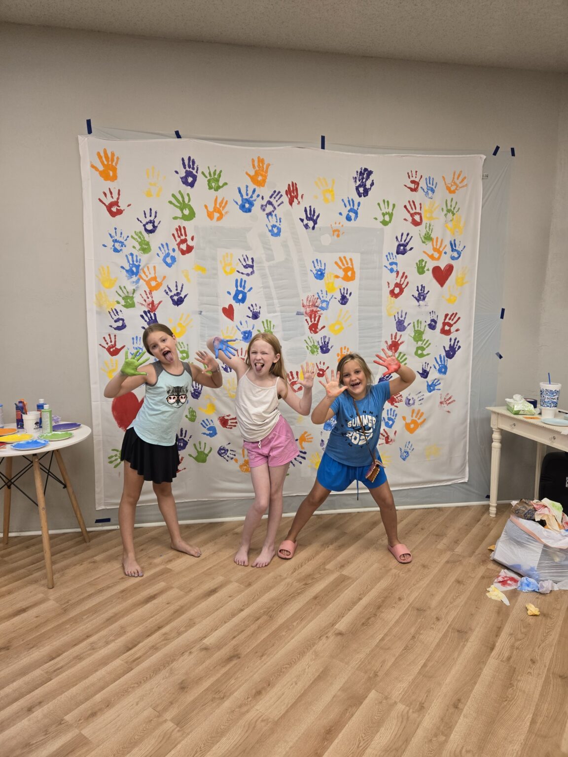 Three children with painted hands in front of a handprint banner during an Inspirations Academy art class